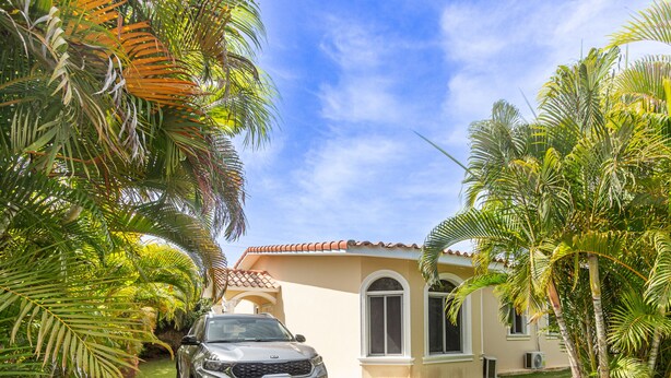A house with a car parked in front, surrounded by palm trees and a clear sky. - Sosua Ocean Village, villa 19a (Sosúa)