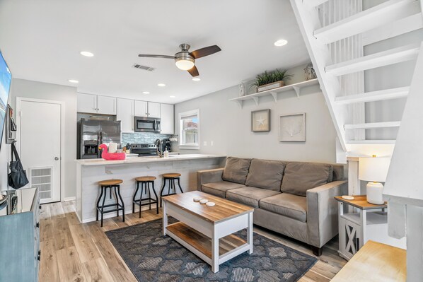 Eine moderne Küche mit weißen Schränken, einer Mikrowelle und einem Kühlschrank. Es gibt einen Holztisch mit Hockern, ein graues Sofa und eine Treppe mit weißen Geländern.