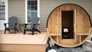 Eine Saunakabine aus Holz mit einer Sitzbank im Inneren, auf einer Terrasse mit zwei Adirondack-Stühlen und einem kleinen Tisch.