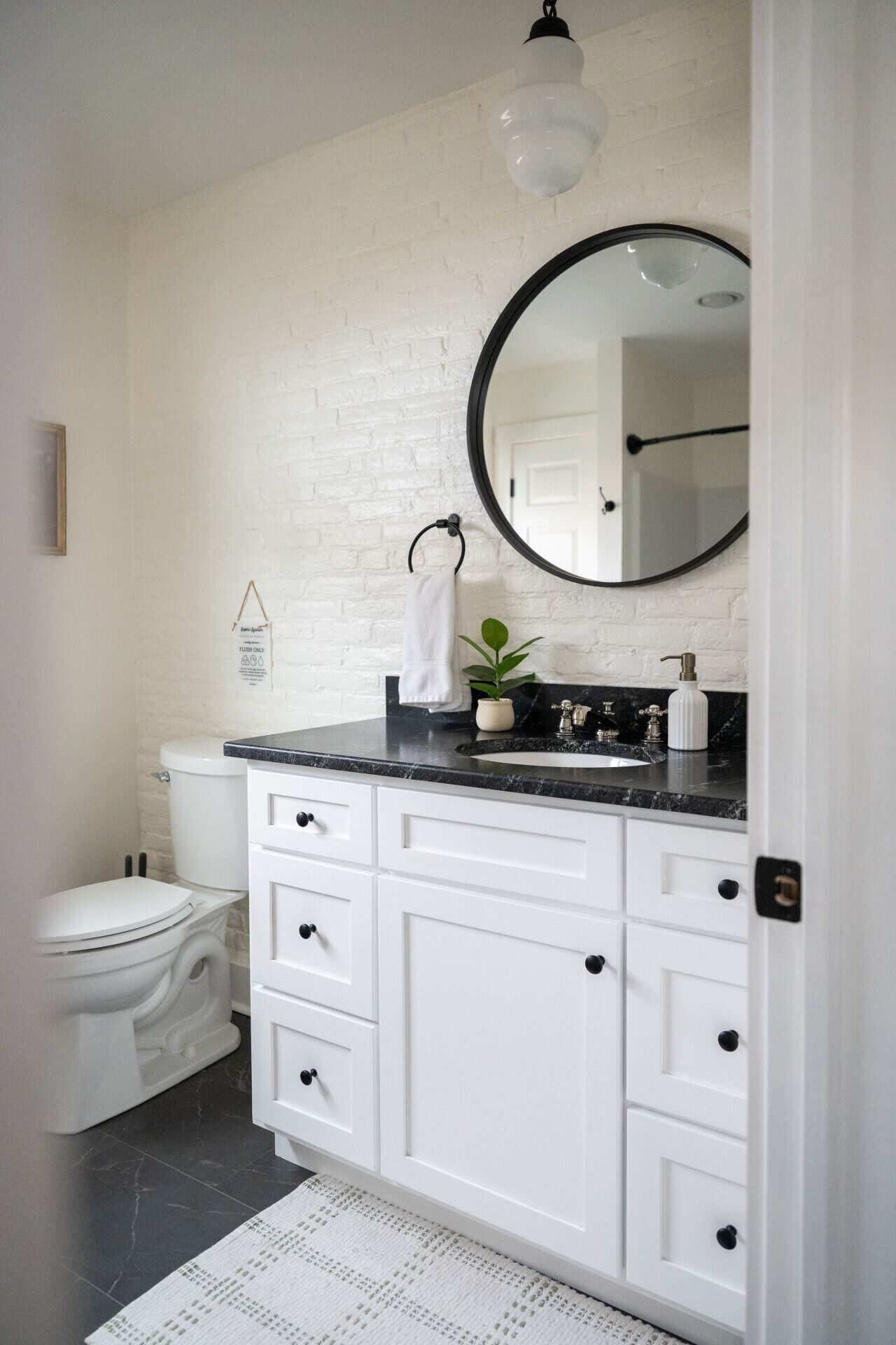 Un bagno con un water bianco, un lavabo su un piano nero, un mobile vanità bianco con cassetti, uno specchio rotondo e una lampada a sospensione.