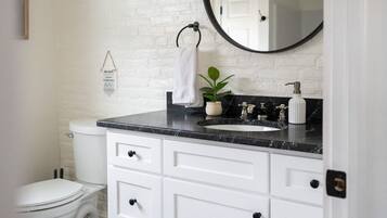 Un bagno con un water bianco, un lavabo su un piano nero, un mobile vanità bianco con cassetti, uno specchio rotondo e una lampada a sospensione.