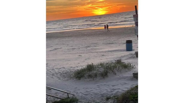 Une scène de plage au coucher du soleil, avec deux personnes qui se promènent et une poubelle.
