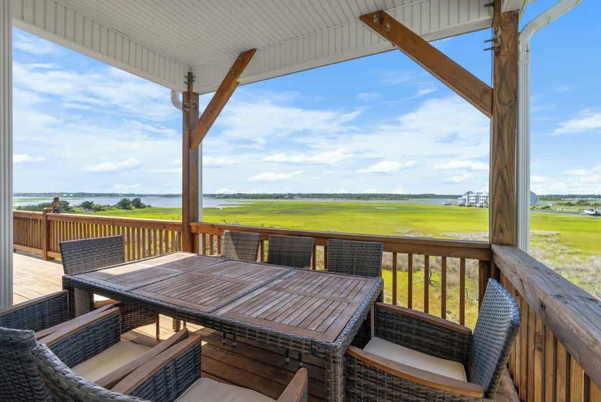 A covered outdoor area with a table and chairs, a wooden railing, and a view of a green landscape.