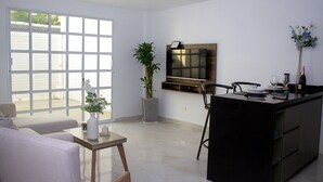 A modern living room with a sofa, a coffee table, a TV mounted on the wall, a potted plant, and a dining area with chairs and a table.