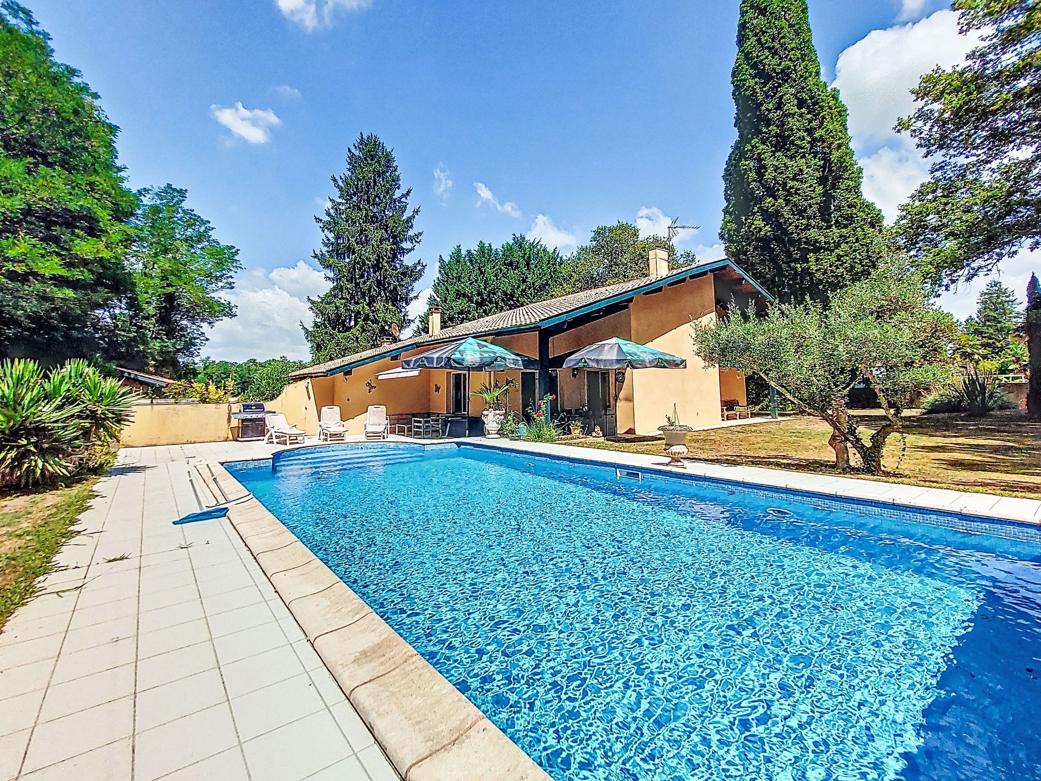 En swimmingpool med en kakelbeklädd poolkant, en täckt uteplats med stolar och ett hus i bakgrunden.