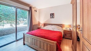 A bedroom with a wooden bed, a nightstand with a lamp, a wardrobe, and a sliding glass door leading to an outdoor area. - Maison du Chêne Pointu by Interhome (Saint Pandelon)