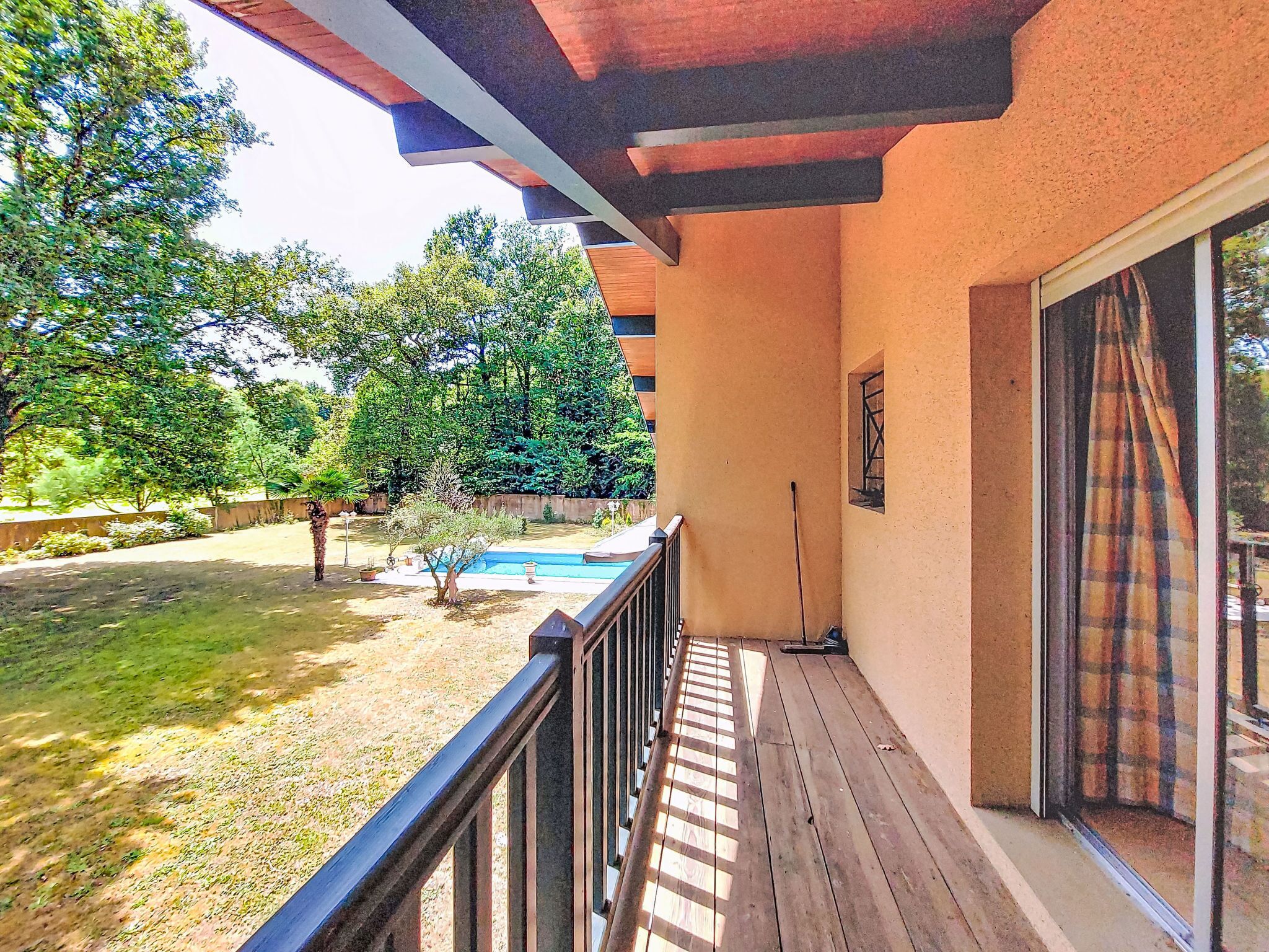 A covered outdoor area with a wooden deck, a small pool, and a view of a garden with trees and shrubs.