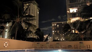 A nighttime pool area with clear water, palm trees, and illuminated buildings in the background. - Newly renovated, secure, 2-bedroom apartment in beautiful Waikiki with AC (Honolulu)