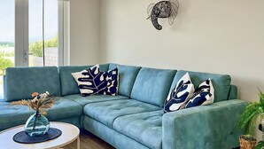 A teal sectional sofa with abstract patterned pillows, a small round table with a vase, and a potted plant. - 2 bedroom accommodation in Wieringerwerf (Wieringerwerf)