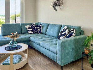 Un sofá seccional color turquesa con almohadones de diseño abstracto, una mesita redonda con un florero y una planta en maceta.