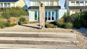 Une propriété située en bord de mer, avec une promenade en bois menant à une maison bleu clair dotée d’un toit foncé.