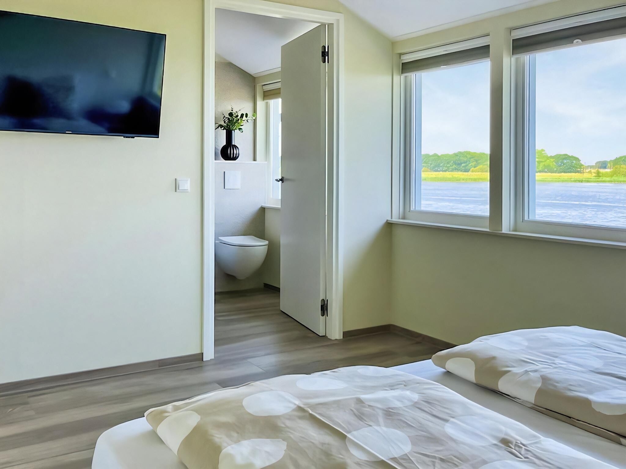 Une chambre à coucher avec un lit, une télévision, une fenêtre donnant sur un paysage verdoyant, et une salle de bain visible à travers une porte ouverte.