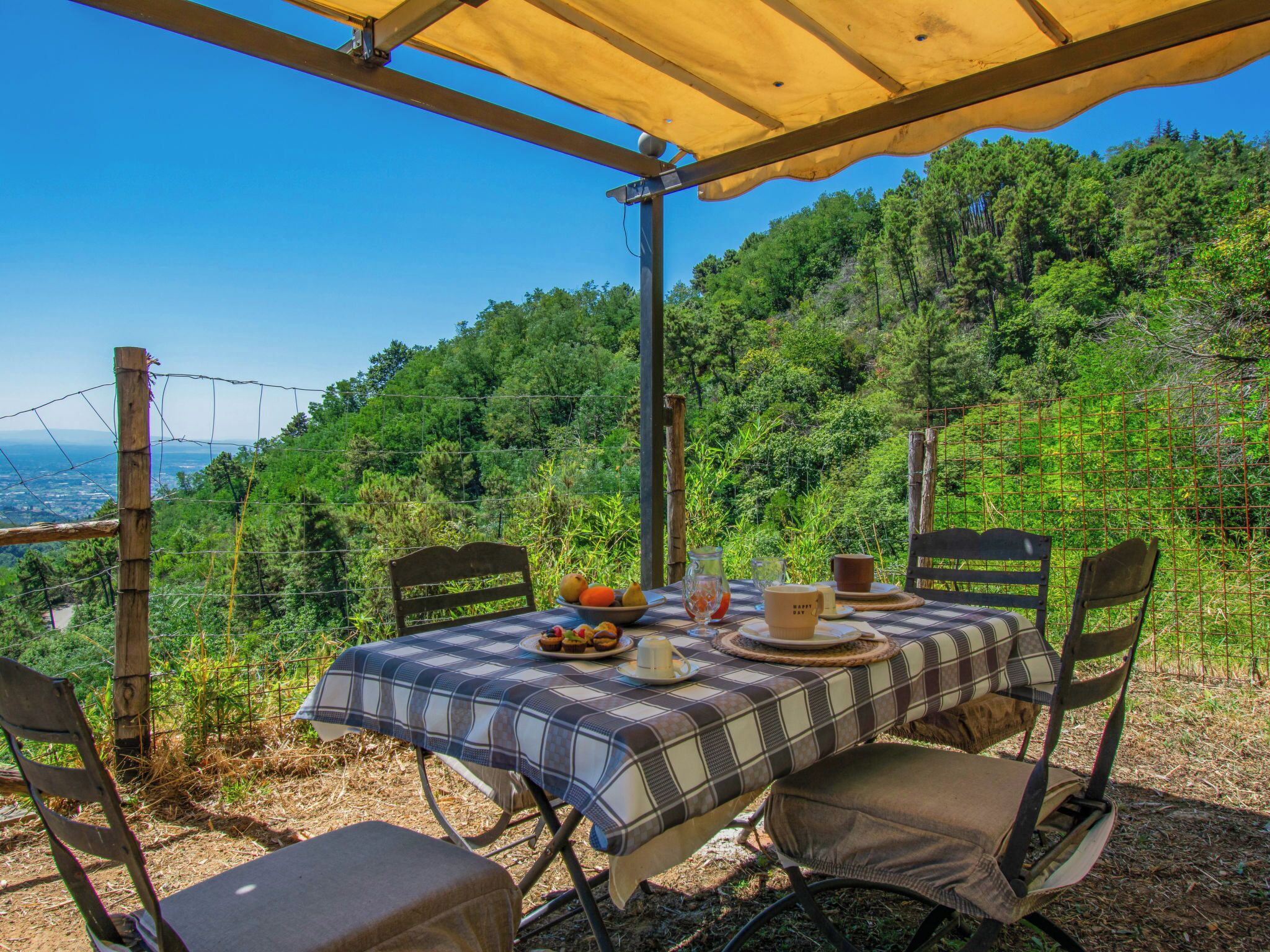 An outdoor dining area with a table set for two, surrounded by chairs, overlooking a forested hillside.