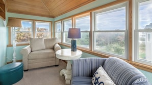 Une pièce chaleureuse avec un plafond en bois, de grandes fenêtres, un canapé beige, un fauteuil à rayures et une petite table blanche sur laquelle se trouve une lampe.