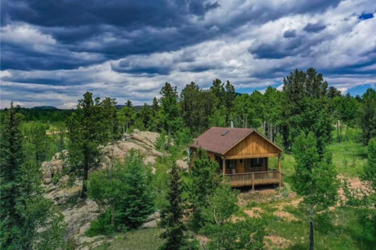 A wooden cabin with a balcony, surrounded by dense forest and a rocky landscape under a cloudy sky.