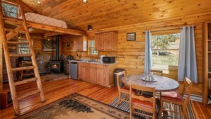 A wooden cabin interior with a loft bedroom, a kitchen with wooden cabinets, a dining area with a round table, and a stone fireplace. - Phantom Canyon Resort (Victor)