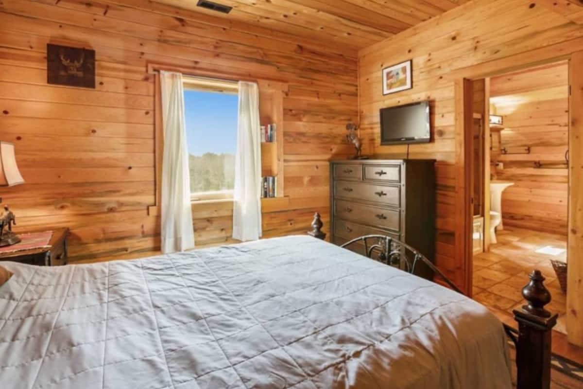 A wooden cabin bedroom with a bed, dresser, television, and a window with curtains.