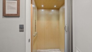 A narrow hallway with a wooden door, a metal call button, and a framed notice on the wall. - Classic 4BR Cherry Grove home w/ elevator, full kitchen & short walk to beach (North Myrtle Beach)