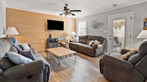 A living room with a wooden wall, a ceiling fan, a flat-screen TV, and a sliding glass door to the outside. - Classic 4BR Cherry Grove home w/ elevator, full kitchen & short walk to beach (North Myrtle Beach)