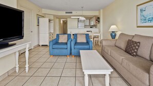 Un salon avec un téléviseur, deux fauteuils bleus, un canapé beige et une table basse blanche.