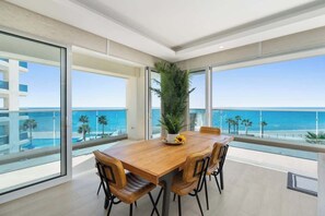 Un comedor moderno con una mesa y sillas de madera, ventanas amplias que ofrecen vista al océano y un edificio al otro lado del agua.