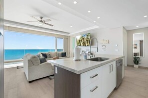 A modern kitchen with a central island, a built-in dishwasher, and a large window overlooking the sea.