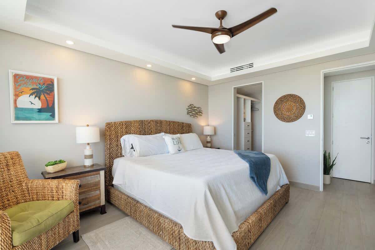 Une chambre avec un grand lit, un ventilateur de plafond, une table de chevet, un fauteuil en osier et un tableau accroché au mur.