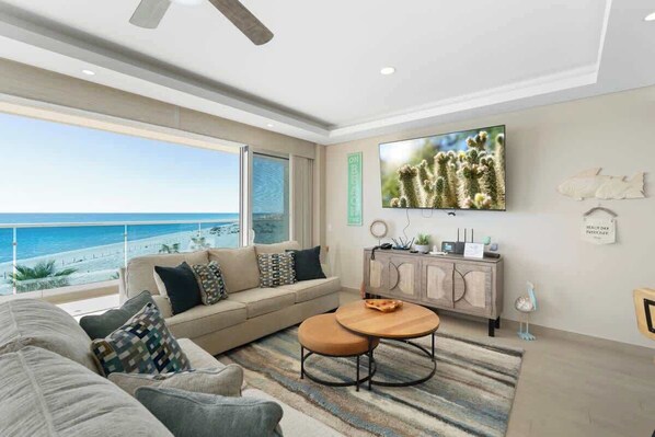 A living room with a large window overlooking a beach, a sofa, a coffee table, and a flat-screen TV mounted on the wall.