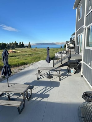 A patio with picnic tables and umbrellas overlooking a lake, with a building adjacent to the patio. - Sleeps 30 • Hot Tub • Sauna • Pool • Walk to Bear Lake Marina (Garden City)