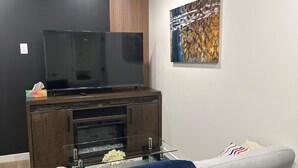 A modern living room with a flat-screen TV on a wooden entertainment unit, a glass coffee table, a grey sofa, and a blue throw blanket. - Restful Haven (Winnipeg)