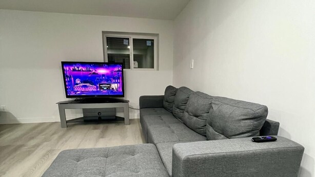 Un salon moderne avec un canapé d’angle gris, un pouf assorti et un téléviseur à écran plat posé sur un meuble.