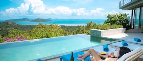 Piscina privada infinita con vistas al océano y montañas, sillas de descanso y una mesa de comedor con comida y bebidas.