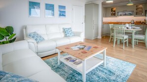 A modern living room with a white sofa, a wooden coffee table with magazines, a blue patterned rug, and a kitchen area with a dining table and chairs.