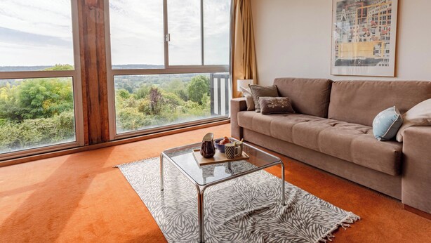 A living room with a large window, a brown sofa, a glass coffee table, and a patterned rug. - La Pinchonnière by Interhome (Deauville-Trouville)