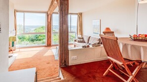 A spacious room with a large window, a wooden chair, a table with a basket of fruit, and a sofa with cushions. - La Pinchonnière by Interhome (Deauville-Trouville)