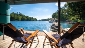 Une terrasse couverte avec deux chaises longues donnant sur un lac, un quai avec un bateau, et un belvédère en arrière-plan.