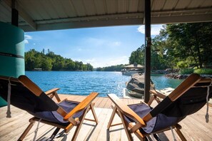Una terraza techada con dos sillones de relax que dan a un lago, un muelle con un bote, y un pabellón al fondo.