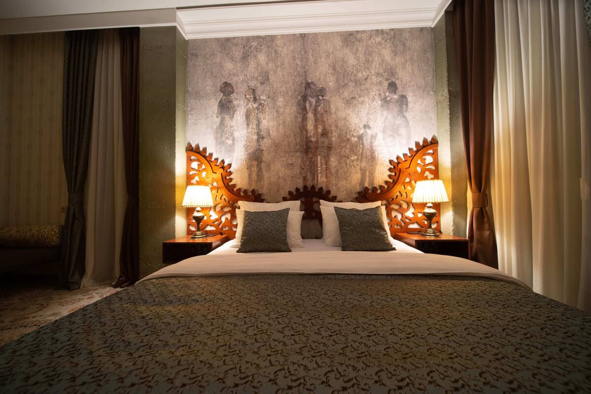 Una habitación de hotel con una cama grande, dos mesitas de noche con lámparas y un mural texturizado en la pared.