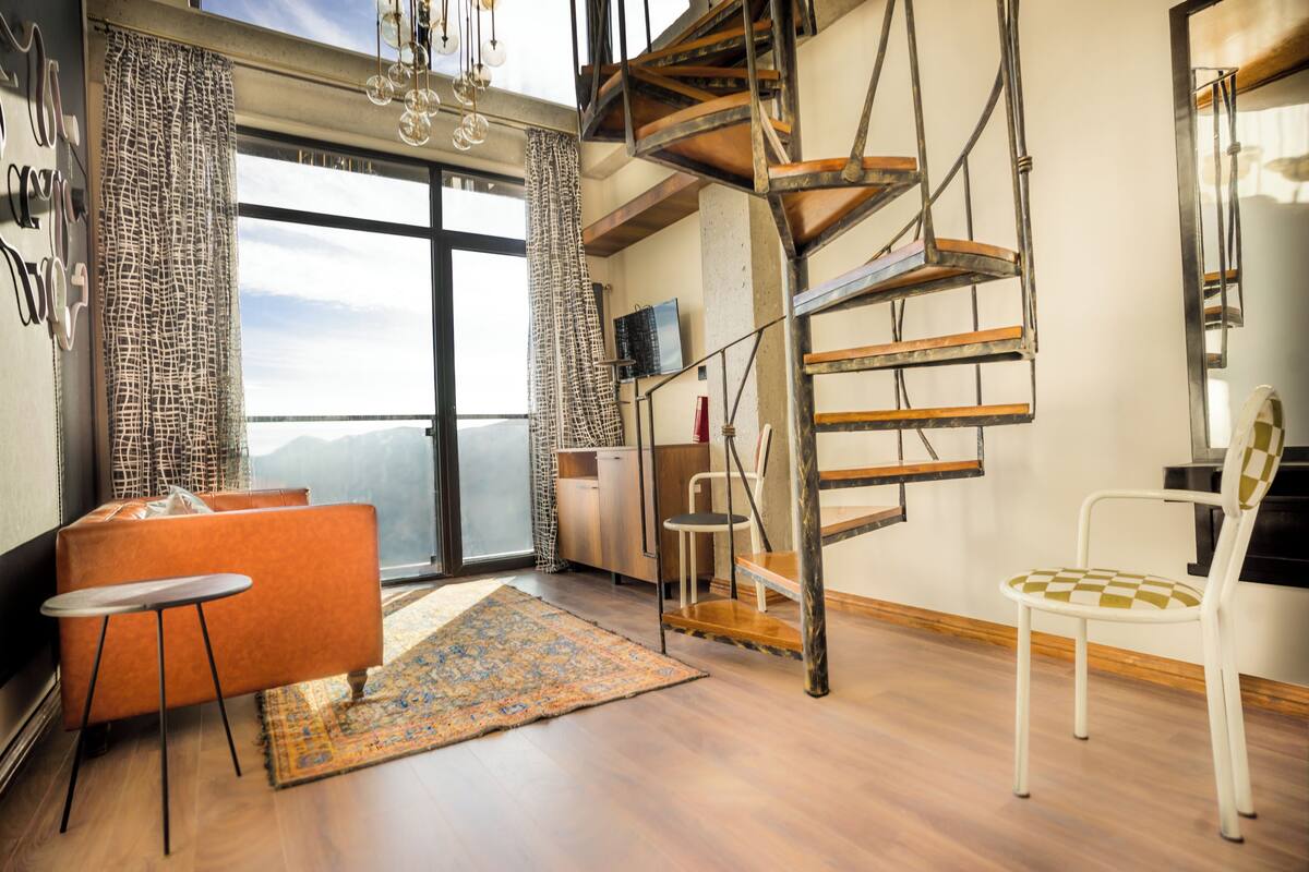 Una sala de estar moderna con una escalera de caracol, un sofá, una mesita y vistas a las montañas.