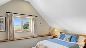 Een slaapkamer met schuine wand, een raam met gordijnen, een nachtkastje met lamp, een bed met blauw beddengoed en een klein houten krukje.