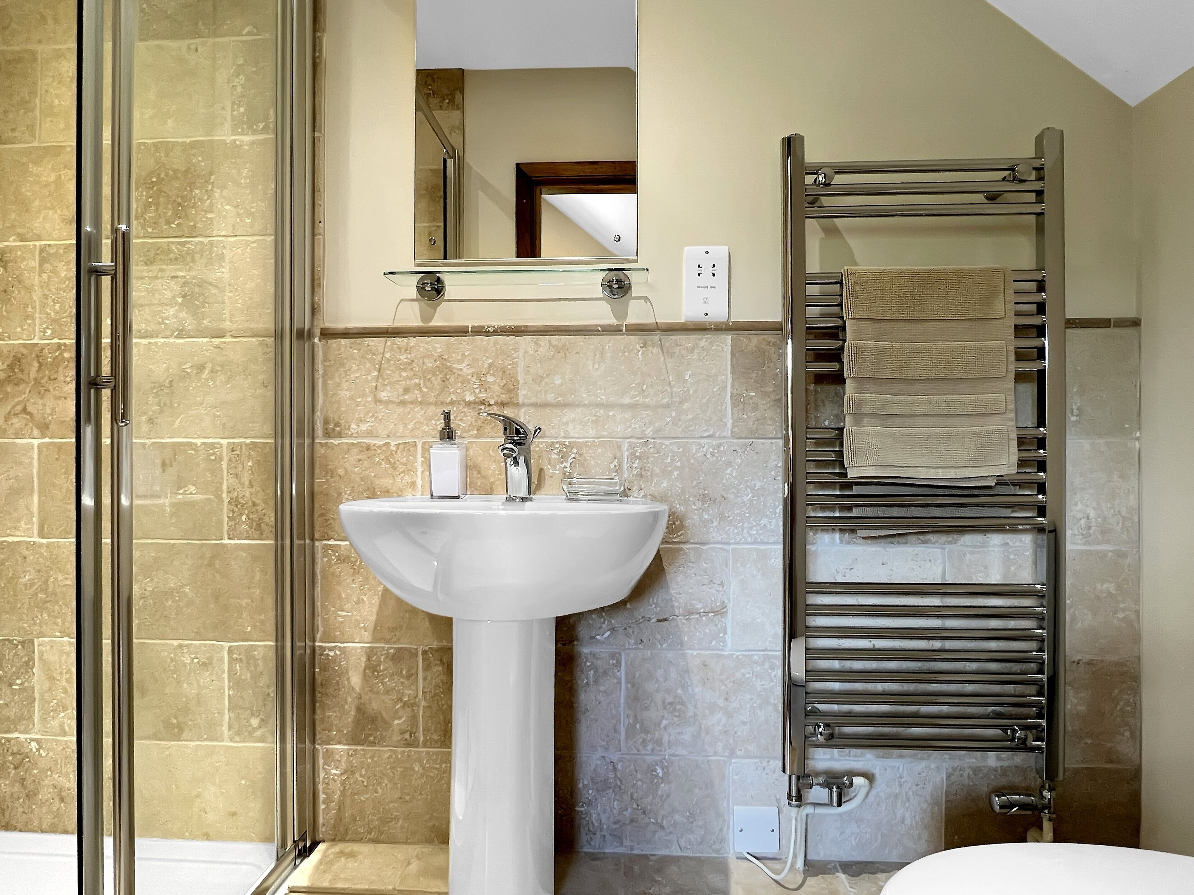 Bagno con box doccia in vetro, lavandino bianco su piedistallo, porta asciugamani e specchio sopra il lavandino.