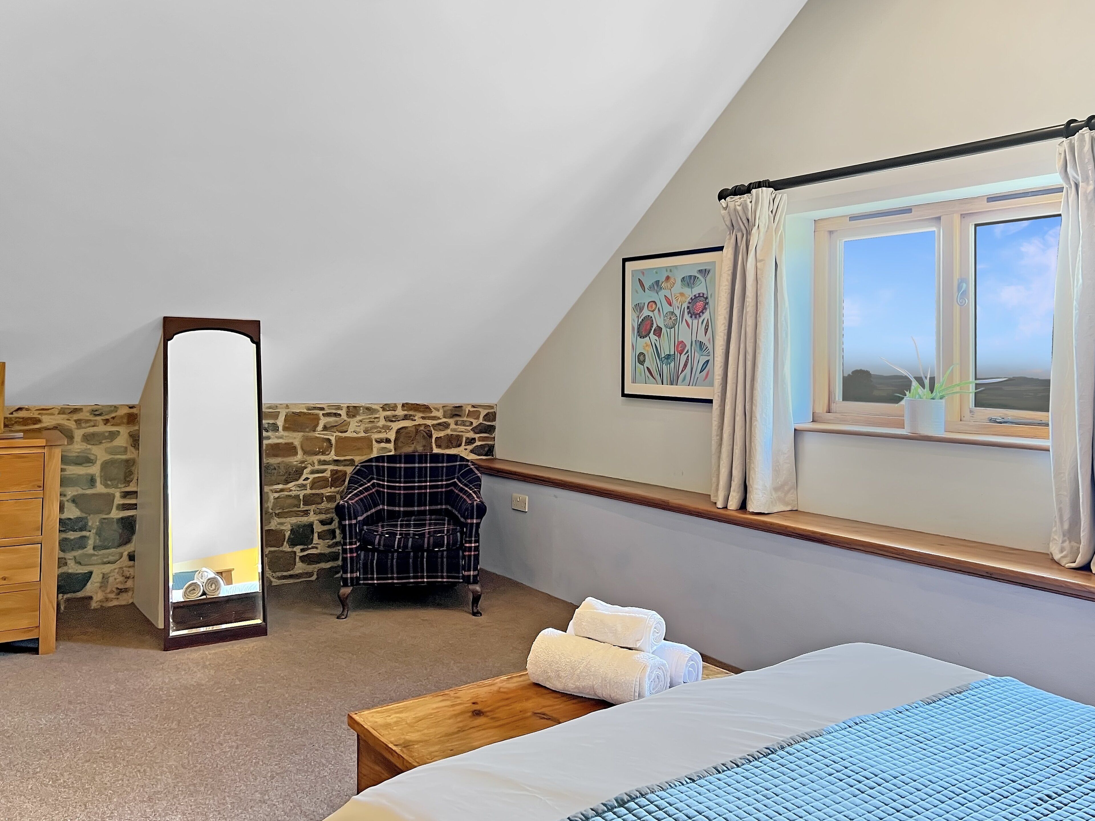 Une chambre à coucher avec un lit, une commode en bois, un miroir, un mur en pierre, une fenêtre avec des rideaux et un tableau.
