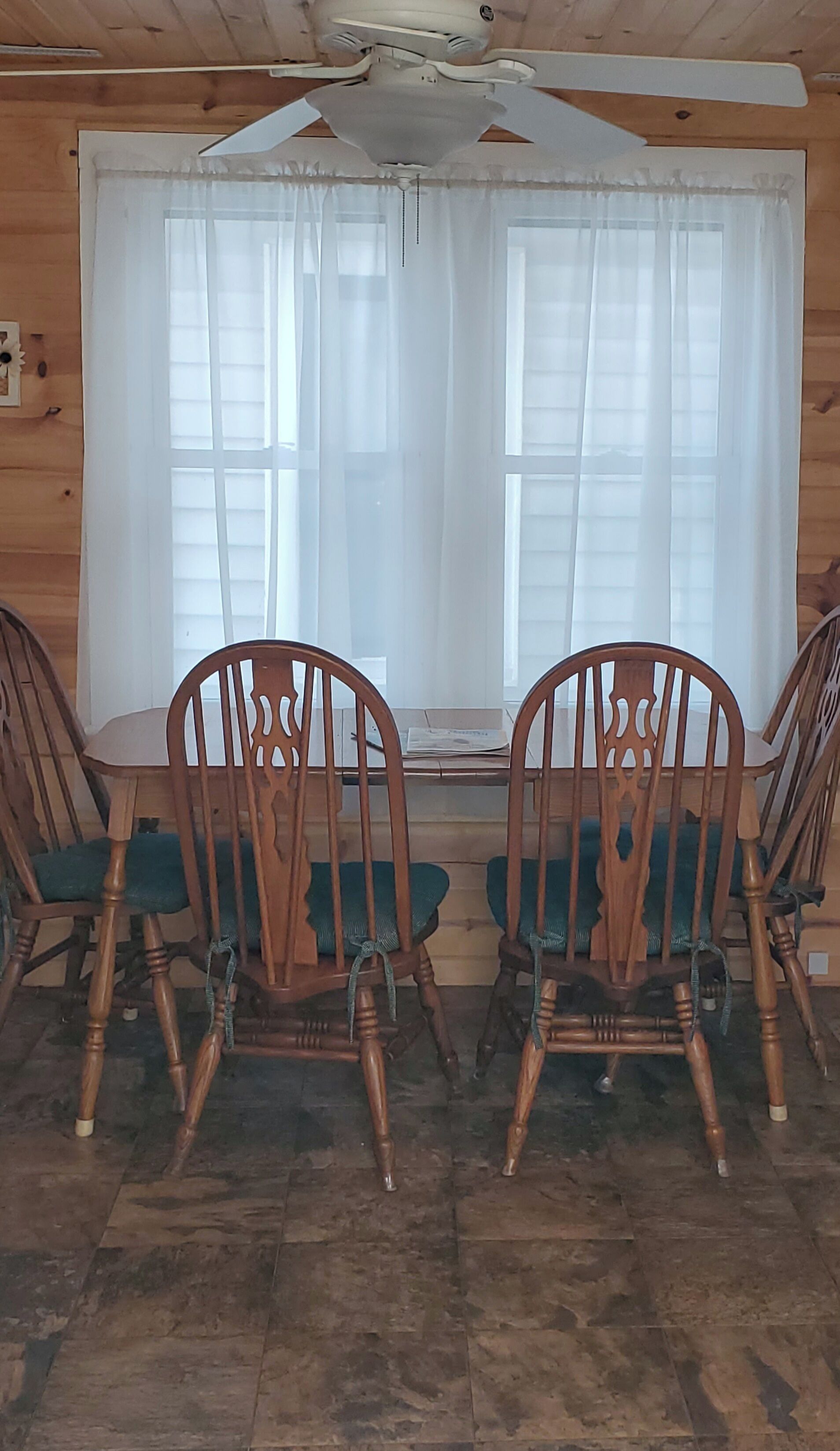 A wooden interior with two wooden chairs with green cushions, a wooden table, and a window with white curtains.