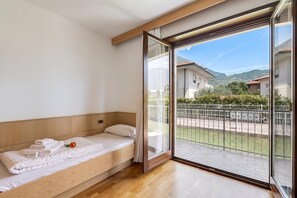 A hotel room with a wooden bed, a large sliding glass door leading to a balcony, and a view of the surrounding buildings and hills.