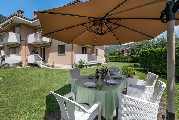 A dining area with a table set for four, surrounded by white wicker chairs, under a large beige umbrella.