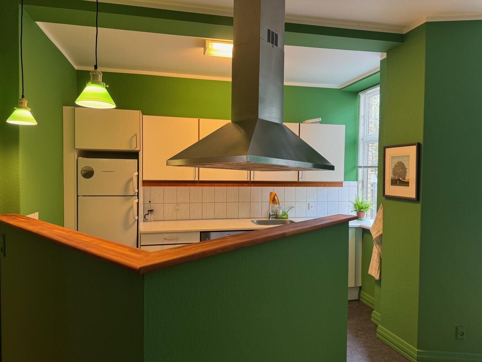 A kitchen with green walls, white cabinets, and a wooden countertop. There is a stainless steel range hood above the stove, and pendant lights hanging above.