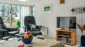 Ein Wohnzimmer mit einer Ledersofa, einem Fernseher auf einem Holzschrank, einem Tisch mit Obstschale und einem Fenster mit Vorhängen.