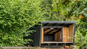 Eine kleine Holzhütte mit offener Vorderseite, umgeben von üppigem Grün.