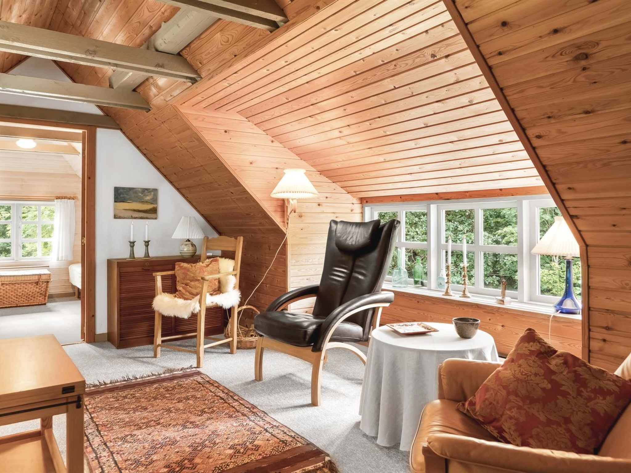 A cozy attic room with wooden beams, a black leather chair, a wooden sofa, a small round table, and a view of greenery through large windows.