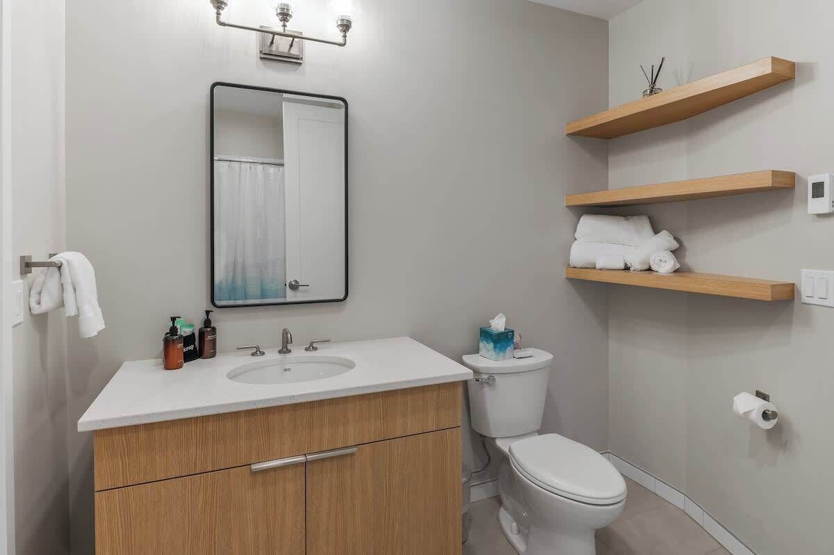 Une salle de bain moderne avec un lavabo blanc, des meubles en bois, des toilettes et des étagères en bois au-dessus des toilettes.